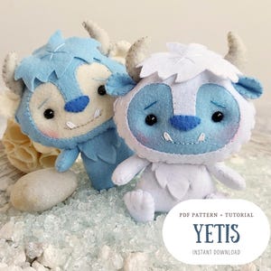 May include: Two plush Yeti toys, one blue and one white, with embroidered facial features. The toys are sitting on a bed of white and blue decorative stones. The text "PDF PATTERN + TUTORIAL YETIS INSTANT DOWNLOAD" is at the bottom of the image.