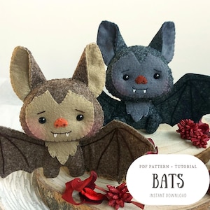 Pode incluir: Dois brinquedos de peluche de morcego de feltro com detalhes costurados. O morcego à esquerda é castanho com uma cara castanho claro e o morcego à direita é cinzento com uma cara cinzento claro. Ambos os morcegos têm asas pretas e estão sentados numa superfície de madeira com acentos de flores vermelhas. O texto "PDF PATTERN + TUTORIAL BATS INSTANT DOWNLOAD" está abaixo dos morcegos.