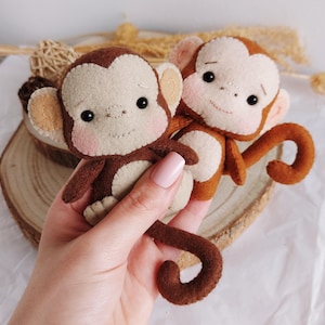 Può includere: Due bambole a forma di scimmia in feltro fatte a mano. Le scimmie hanno corpi marroni, facce color camoscio e guance rosa. Hanno piccoli occhi neri e sorridono. Una scimmia tiene l'altra. Le bambole sono su una fetta di legno.