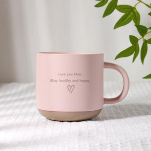 Puede incluir: Una taza de cerámica rosa con base marrón claro y asa curva. La taza tiene el texto "Love you Mum, Stay healthy and happy." con un símbolo de corazón. La taza está sobre una superficie blanca.