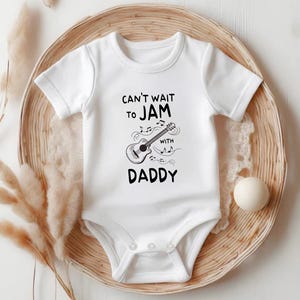 May include: White baby onesie with short sleeves and a black graphic of a guitar with musical notes and the text "Can't wait to jam with Daddy".