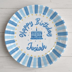 May include: A round, white ceramic plate with a blue and light blue striped border. The plate features the words "Happy Birthday Josiah" in blue script, along with a blue birthday cake with a lit candle. Small gold hearts are scattered around the edge.
