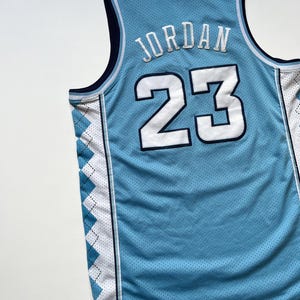 May include: Light blue basketball jersey with the name "JORDAN" and the number "23" in white. The jersey has a mesh texture and features navy blue and white accents, including a diamond pattern on the sides.
