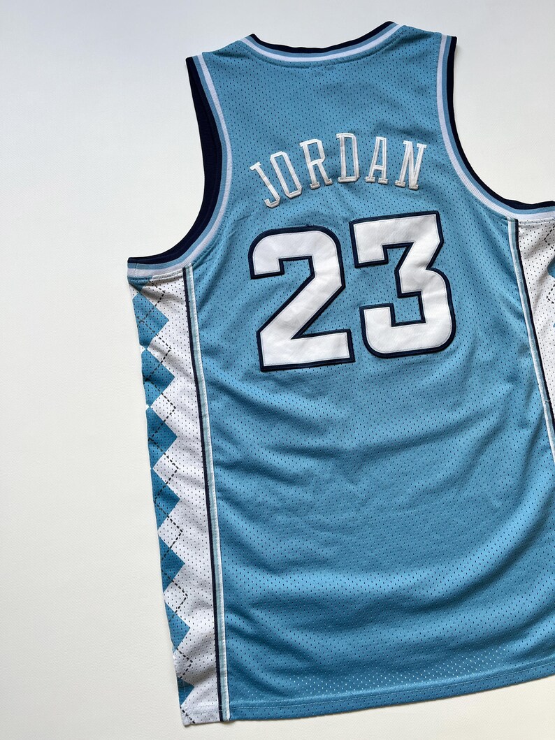 May include: Light blue basketball jersey with the name "JORDAN" and the number "23" in white. The jersey has a mesh texture and features navy blue and white accents, including a diamond pattern on the sides.