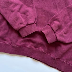 Puede incluir: Primer plano de una sudadera burdeos. La imagen destaca los pu&ntilde;os y el dobladillo acanalados de la prenda. La tela parece suave y ligeramente desgastada, con un enfoque en la textura y el color del material. El fondo es una superficie blanca lisa.