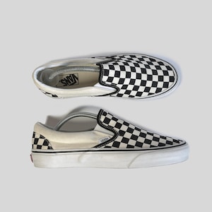 Könnte beinhalten: Zwei schwarz-weiß karierte Vans Slip-On-Sneaker. Die Schuhe haben eine weiße Sohle und ein schwarzes Gummiband oben. Das Vans-Logo ist auf der Innensohle und der Ferse sichtbar.