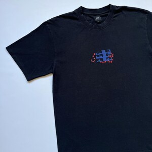 K&ouml;nnte beinhalten: Schwarzes T-Shirt mit kurzen &Auml;rmeln und Rundhalsausschnitt. Das Shirt hat einen rechteckigen Aufdruck auf der Vorderseite mit blauen und roten abstrakten Formen. Aus weichem Material, geeignet f&uuml;r den l&auml;ssigen Gebrauch.