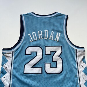 May include: Light blue basketball jersey with the name "JORDAN" and the number "23" in white. The jersey has a mesh design and white and blue argyle pattern on the sides. The neckline and armholes are trimmed in navy blue.