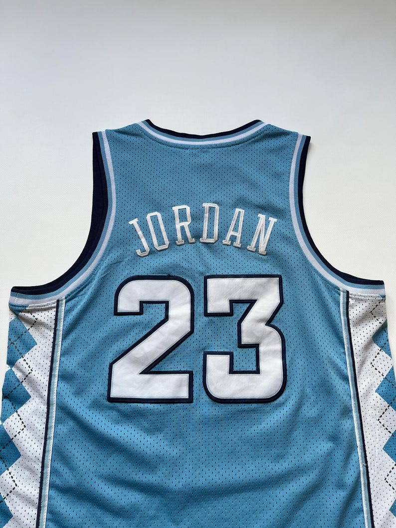 May include: Light blue basketball jersey with the name "JORDAN" and the number "23" in white. The jersey has a mesh design and white and blue argyle pattern on the sides. The neckline and armholes are trimmed in navy blue.