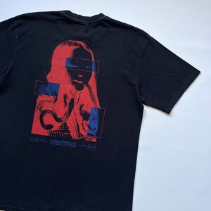 K&ouml;nnte beinhalten: Schwarzes T-Shirt mit einer rot-blauen Grafik eines menschlichen Gesichts und abstrakten Formen. Die Grafik enth&auml;lt den wei&szlig;en Text "SECOND PRINCESS". Das T-Shirt ist aus weichem Material.
