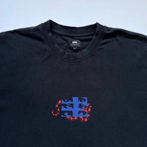 K&ouml;nnte beinhalten: Schwarzes T-Shirt mit Rundhalsausschnitt und Grafikdesign. Das Design zeigt ein blaues Quadrat mit mehreren Augen und eine rote schlangenartige Form. Der Markenname "EDWIN" ist auf der Brust aufgedruckt.