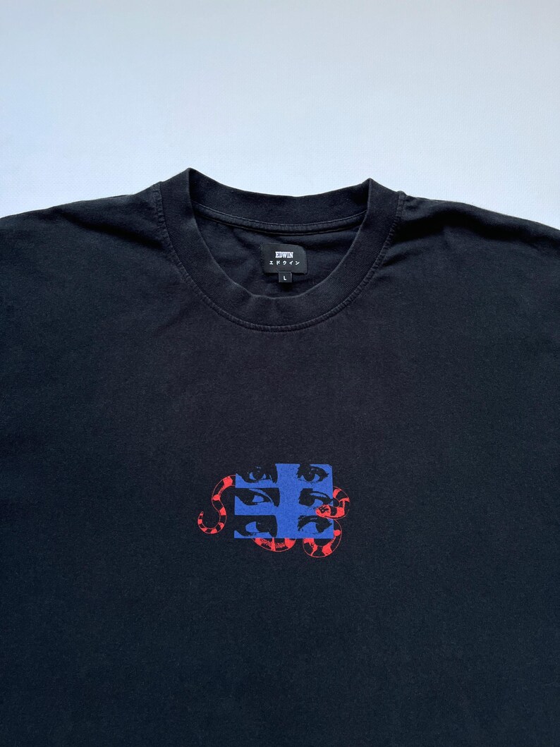 K&ouml;nnte beinhalten: Schwarzes T-Shirt mit Rundhalsausschnitt und Grafikdesign. Das Design zeigt ein blaues Quadrat mit mehreren Augen und eine rote schlangenartige Form. Der Markenname "EDWIN" ist auf der Brust aufgedruckt.