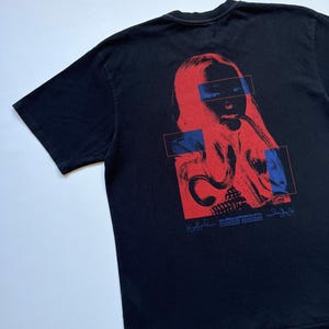 K&ouml;nnte beinhalten: Schwarzes T-Shirt mit einer rot-blauen Grafik eines Gesichts. Das Design umfasst rechteckige blaue Akzente und Text am unteren Rand. Das T-Shirt ist aus weichem Material.