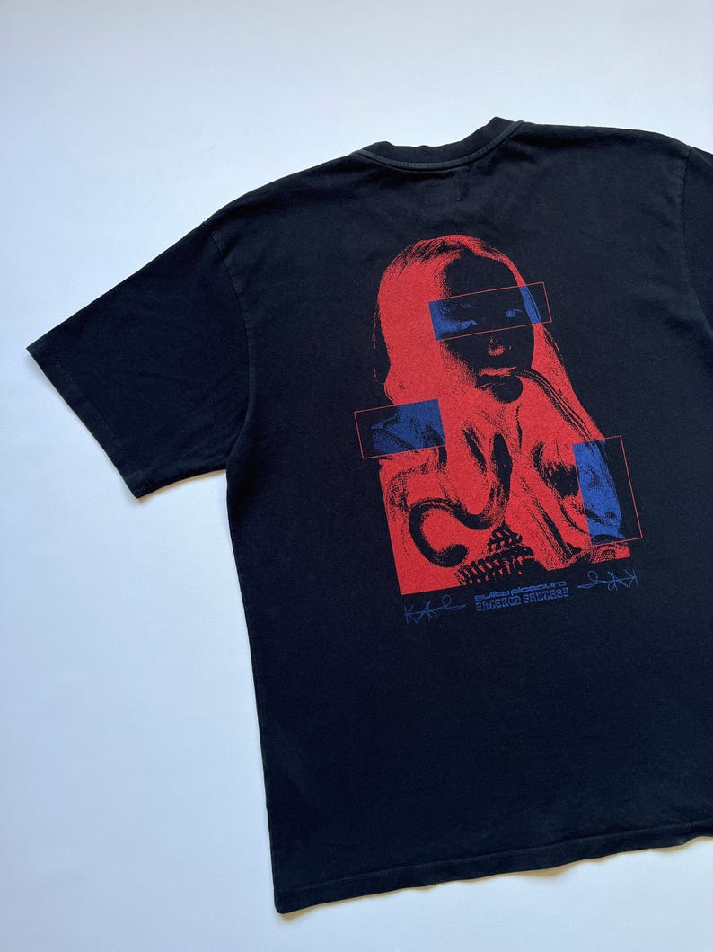 K&ouml;nnte beinhalten: Schwarzes T-Shirt mit einer rot-blauen Grafik eines Gesichts. Das Design umfasst rechteckige blaue Akzente und Text am unteren Rand. Das T-Shirt ist aus weichem Material.