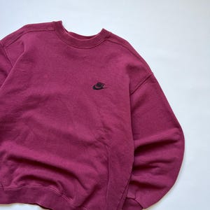 Puede incluir: Una sudadera burdeos de cuello redondo con un peque&ntilde;o logotipo negro de Nike en el pecho. La sudadera est&aacute; hecha de una tela suave y texturizada y tiene un ajuste cl&aacute;sico y relajado. El fondo es blanco liso.