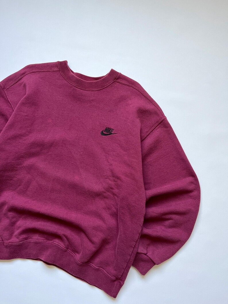 Puede incluir: Una sudadera burdeos de cuello redondo con un peque&ntilde;o logotipo negro de Nike en el pecho. La sudadera est&aacute; hecha de una tela suave y texturizada y tiene un ajuste cl&aacute;sico y relajado. El fondo es blanco liso.