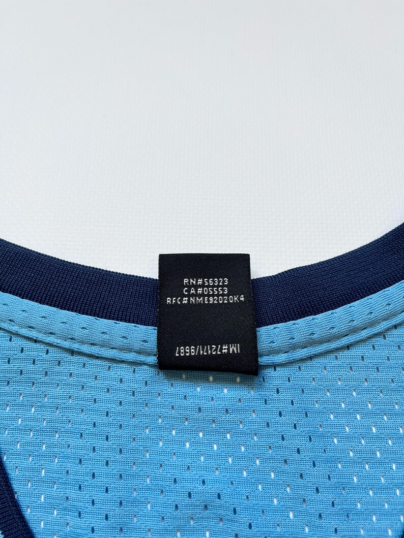 May include: Close-up of a light blue mesh jersey with a navy blue collar. A black tag displays text including "RN#56323" and "CA#05553". The jersey has a perforated design.