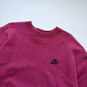 Puede incluir: Una sudadera burdeos de cuello redondo con el logotipo de Nike bordado en negro en la parte superior izquierda del pecho. La sudadera tiene cuello, pu&ntilde;os y dobladillo de canal&eacute;. La tela parece suave y ligeramente descolorida.