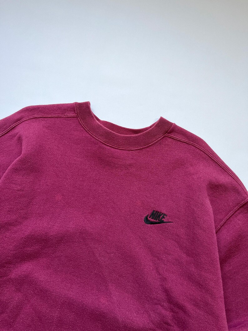 Puede incluir: Una sudadera burdeos de cuello redondo con el logotipo de Nike bordado en negro en la parte superior izquierda del pecho. La sudadera tiene cuello, pu&ntilde;os y dobladillo de canal&eacute;. La tela parece suave y ligeramente descolorida.