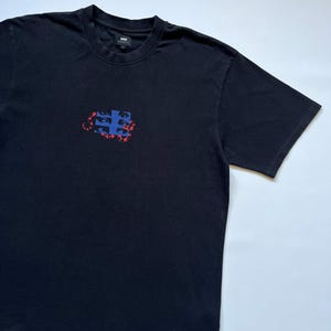 K&ouml;nnte beinhalten: Schwarzes T-Shirt mit Rundhalsausschnitt und kleinem Grafikmuster auf der Brust. Die Grafik ist eine Kombination aus blauen und roten Formen. Das T-Shirt hat kurze &Auml;rmel und eine lockere Passform. Der Markenname ist oben am R&uuml;cken sichtbar.