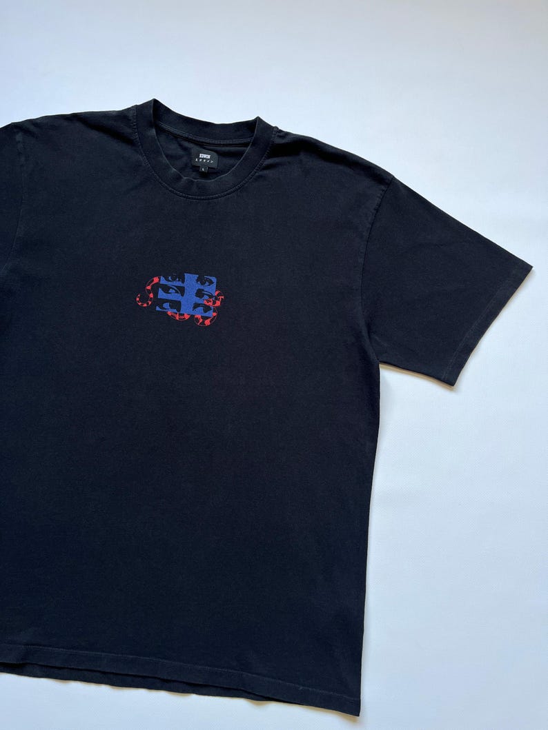 K&ouml;nnte beinhalten: Schwarzes T-Shirt mit Rundhalsausschnitt und kleinem Grafikmuster auf der Brust. Die Grafik ist eine Kombination aus blauen und roten Formen. Das T-Shirt hat kurze &Auml;rmel und eine lockere Passform. Der Markenname ist oben am R&uuml;cken sichtbar.