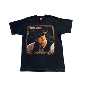 Puede incluir: Camiseta negra con un retrato de Trace Adkins con sombrero de vaquero. La imagen tiene un borde dorado desgastado. El texto "TRACE ADKINS" está encima del retrato y "DREAMIN' OUT LOUD" debajo.