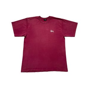 May include: A burgundy t-shirt with short sleeves and a crew neck. It features a small white logo on the left chest. The material looks like a soft cotton blend.