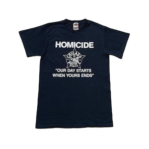 Może przedstawiać: Granatowa koszulka z krótkim rękawem z białym napisem HOMICIDE, grafiką gwiazdy policyjnej z Chicago oraz tekstem OUR DAY STARTS WHEN YOURS ENDS nadrukowanym z przodu. Koszulka posiada okrągły dekolt i standardowy krój.