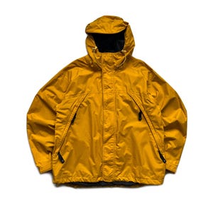 May include: Mustard yellow hooded jacket with a black interior lining. The jacket features a full-length zipper, two front pockets with black zippers, and a drawstring hood. The jacket is designed for outdoor wear and protection from the elements.
