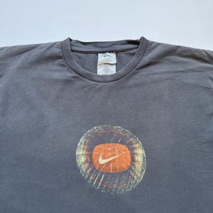 Puede incluir: Camiseta gris con un dise&ntilde;o circular con fondo naranja y el logotipo blanco de Nike. El dise&ntilde;o tiene un aspecto desgastado y vintage. La camiseta tiene cuello redondo y mangas cortas.