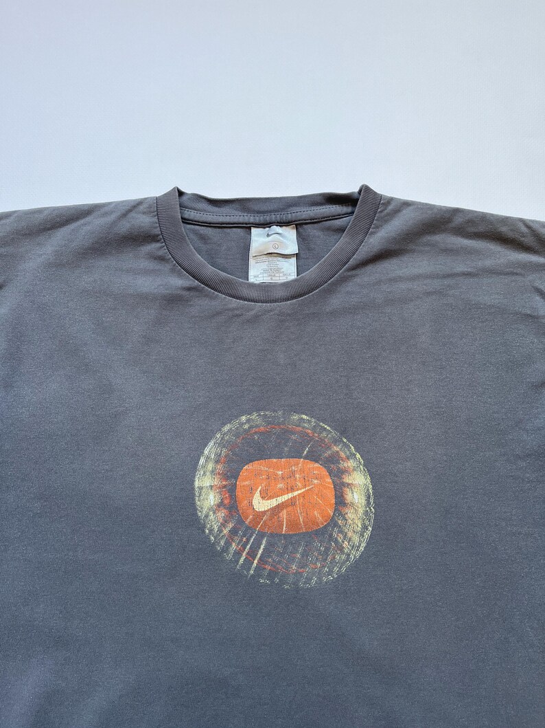 Puede incluir: Camiseta gris con un dise&ntilde;o circular con fondo naranja y el logotipo blanco de Nike. El dise&ntilde;o tiene un aspecto desgastado y vintage. La camiseta tiene cuello redondo y mangas cortas.