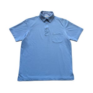 May include: A light blue polo shirt with short sleeves and a chest pocket. The collar features a checkered pattern on the inside. The shirt has three buttons at the placket and a classic fit, suitable for casual wear.