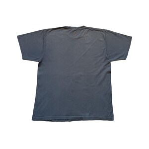 Puede incluir: Una camiseta gris con mangas cortas, colocada plana sobre un fondo blanco. La camiseta tiene cuello redondo y un peque&ntilde;o detalle rojo cerca del escote. La tela parece suave y ligeramente desgastada.