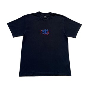 K&ouml;nnte beinhalten: Ein marineblaues T-Shirt mit Rundhalsausschnitt und kurzen &Auml;rmeln. Das Shirt hat ein kleines, abstraktes Design in der Mitte mit blauen und roten Formen. Das T-Shirt ist aus einem weichen, wahrscheinlich Baumwollmaterial.