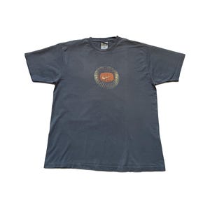 Puede incluir: Camiseta gris oscuro con un dise&ntilde;o gr&aacute;fico circular y el logotipo de Nike en naranja. El logotipo est&aacute; rodeado por un dise&ntilde;o texturizado y descolorido. La camiseta tiene mangas cortas y cuello redondo.