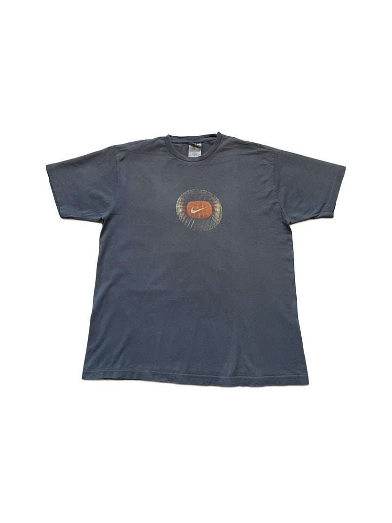 Puede incluir: Camiseta gris oscuro con un dise&ntilde;o gr&aacute;fico circular y el logotipo de Nike en naranja. El logotipo est&aacute; rodeado por un dise&ntilde;o texturizado y descolorido. La camiseta tiene mangas cortas y cuello redondo.