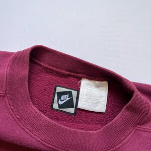Puede incluir: Una sudadera burdeos con cuello redondo y un parche con el logo de Nike. La etiqueta es visible y la sudadera parece estar hecha de un material suave y texturizado. El logo de Nike es un swoosh blanco sobre un cuadrado negro.
