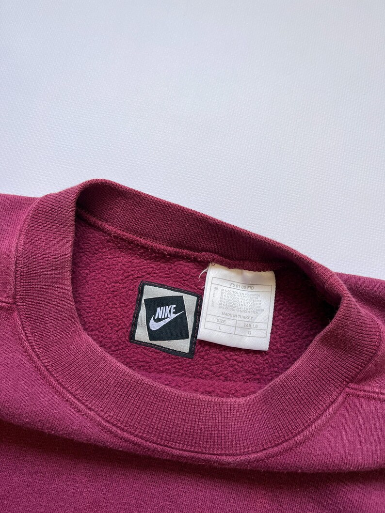 Puede incluir: Una sudadera burdeos con cuello redondo y un parche con el logo de Nike. La etiqueta es visible y la sudadera parece estar hecha de un material suave y texturizado. El logo de Nike es un swoosh blanco sobre un cuadrado negro.