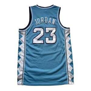 May include: Light blue basketball jersey with the name "JORDAN" and the number "23" in white. The jersey has a mesh design with white and navy blue accents along the sides and neckline. The jersey is sleeveless.