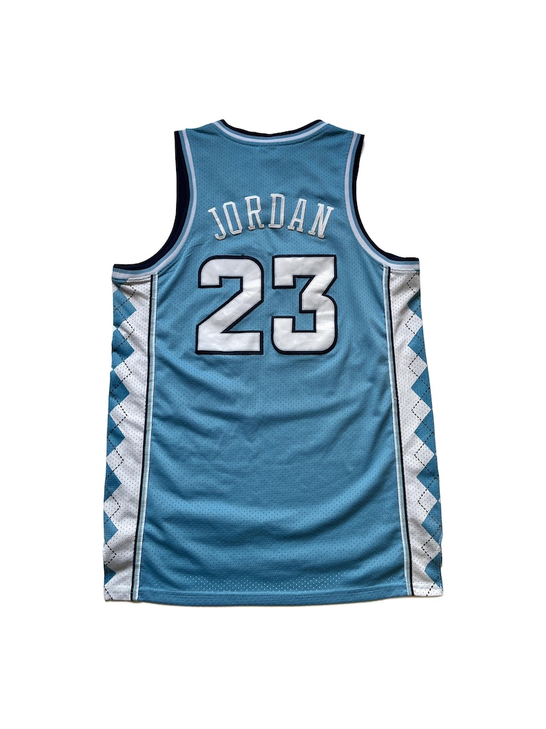 May include: Light blue basketball jersey with the name "JORDAN" and the number "23" in white. The jersey has a mesh design with white and navy blue accents along the sides and neckline. The jersey is sleeveless.