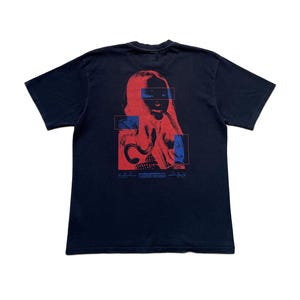 K&ouml;nnte beinhalten: Schwarzes T-Shirt mit einer rot-blauen Grafik eines Frauengesichts. Das Design beinhaltet rechteckige Bl&ouml;cke und Text am unteren Rand. Ein l&auml;ssiges Kleidungsst&uuml;ck.