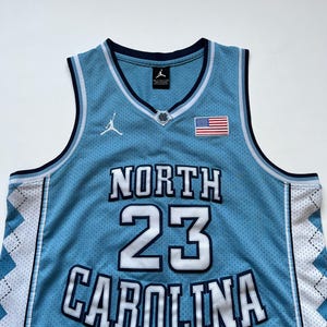 May include: A light blue basketball jersey with the words "NORTH CAROLINA" and the number "23" in white. The jersey has a white and navy blue trim, an American flag patch, and a Jordan logo.