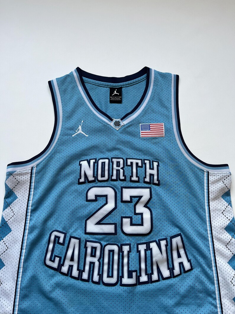 May include: A light blue basketball jersey with the words "NORTH CAROLINA" and the number "23" in white. The jersey has a white and navy blue trim, an American flag patch, and a Jordan logo.