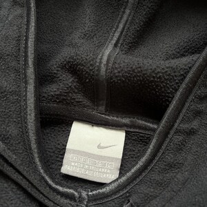 Puede incluir: Primer plano de una sudadera con capucha Nike negra. La capucha est&aacute; levantada y el cord&oacute;n es visible en la parte inferior. Una etiqueta con el logotipo de Nike y texto est&aacute; cosida en la prenda. La tela tiene una apariencia texturizada.