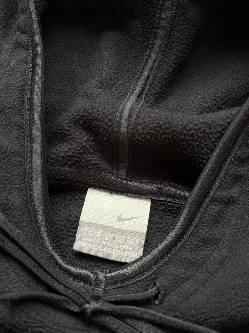 Puede incluir: Primer plano de una sudadera con capucha Nike negra. La capucha est&aacute; levantada y el cord&oacute;n es visible en la parte inferior. Una etiqueta con el logotipo de Nike y texto est&aacute; cosida en la prenda. La tela tiene una apariencia texturizada.