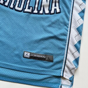 May include: Light blue mesh jersey with the word "CAROLINA" in white block letters with navy blue trim. The jersey has a white and light blue diamond pattern on the side and a "JORDAN" label.