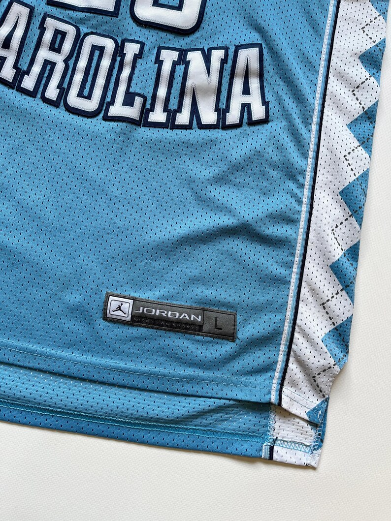May include: Light blue mesh jersey with the word "CAROLINA" in white block letters with navy blue trim. The jersey has a white and light blue diamond pattern on the side and a "JORDAN" label.