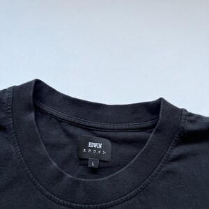 K&ouml;nnte beinhalten: Nahaufnahme eines schwarzen T-Shirts mit Rundhalsausschnitt. Das Shirt hat ein Etikett mit dem Markennamen "EDWIN" in wei&szlig;em Text und japanischen Schriftzeichen. Das Etikett zeigt auch die Gr&ouml;&szlig;e "L" an. Das Shirt ist aus einem dunklen, weichen Stoff gefertigt.