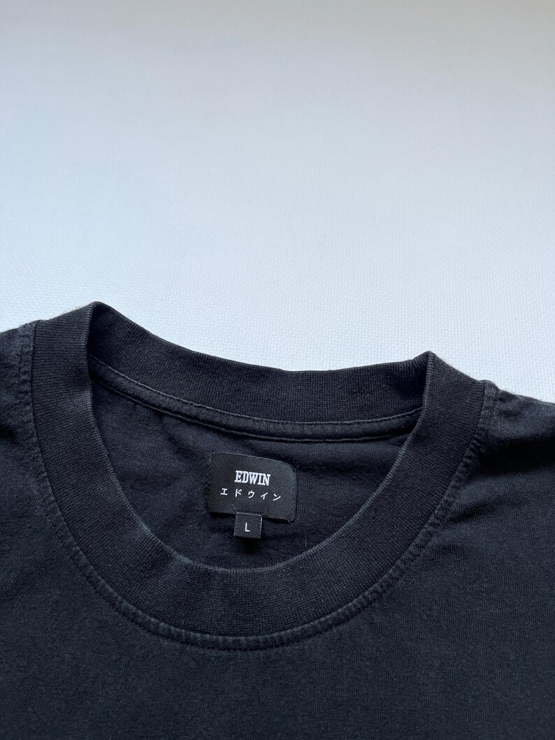 K&ouml;nnte beinhalten: Nahaufnahme eines schwarzen T-Shirts mit Rundhalsausschnitt. Das Shirt hat ein Etikett mit dem Markennamen "EDWIN" in wei&szlig;em Text und japanischen Schriftzeichen. Das Etikett zeigt auch die Gr&ouml;&szlig;e "L" an. Das Shirt ist aus einem dunklen, weichen Stoff gefertigt.