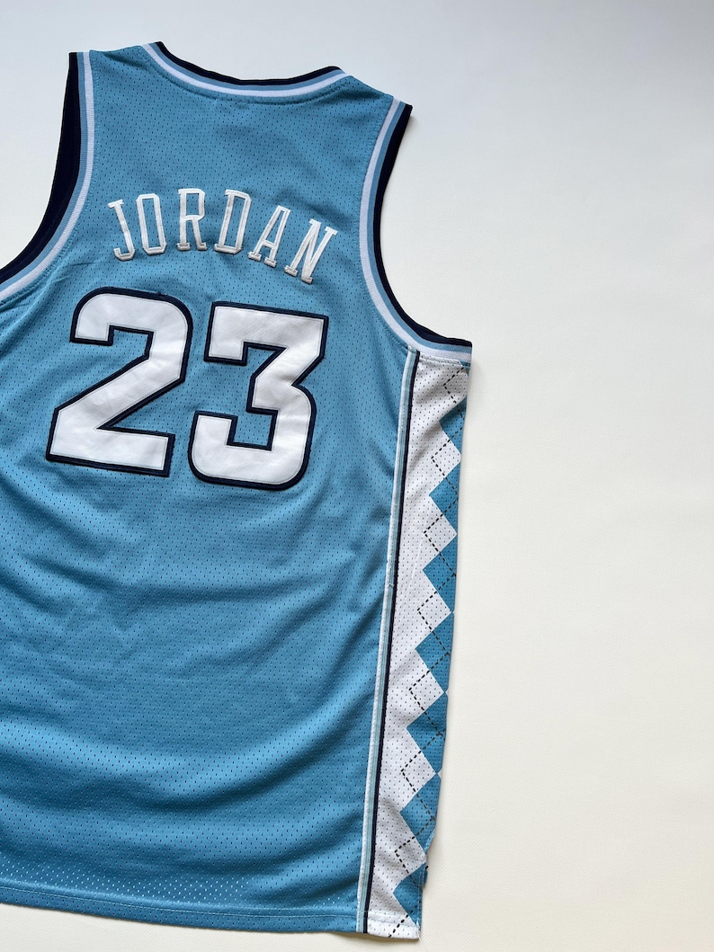 May include: Light blue basketball jersey with the name "JORDAN" and the number "23" in white. The jersey has a mesh design and a white and blue diamond pattern on the side.